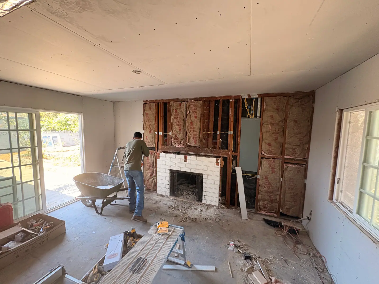 Wide view of a room under renovation with a crew member and a wheelbarrow